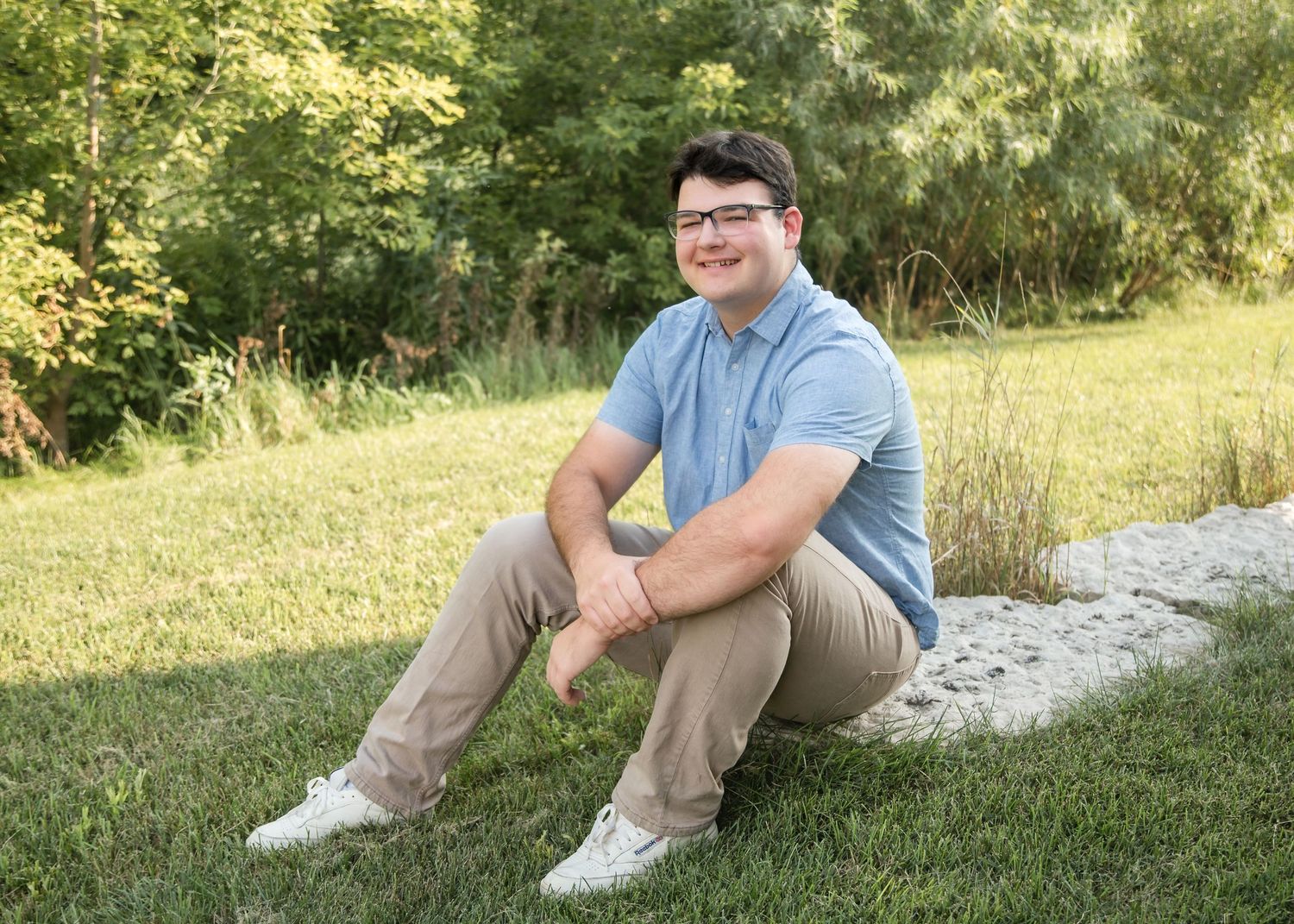 A person in a light blue polo shirt and khaki pants sits casually on a grassy slope on a sunny day.