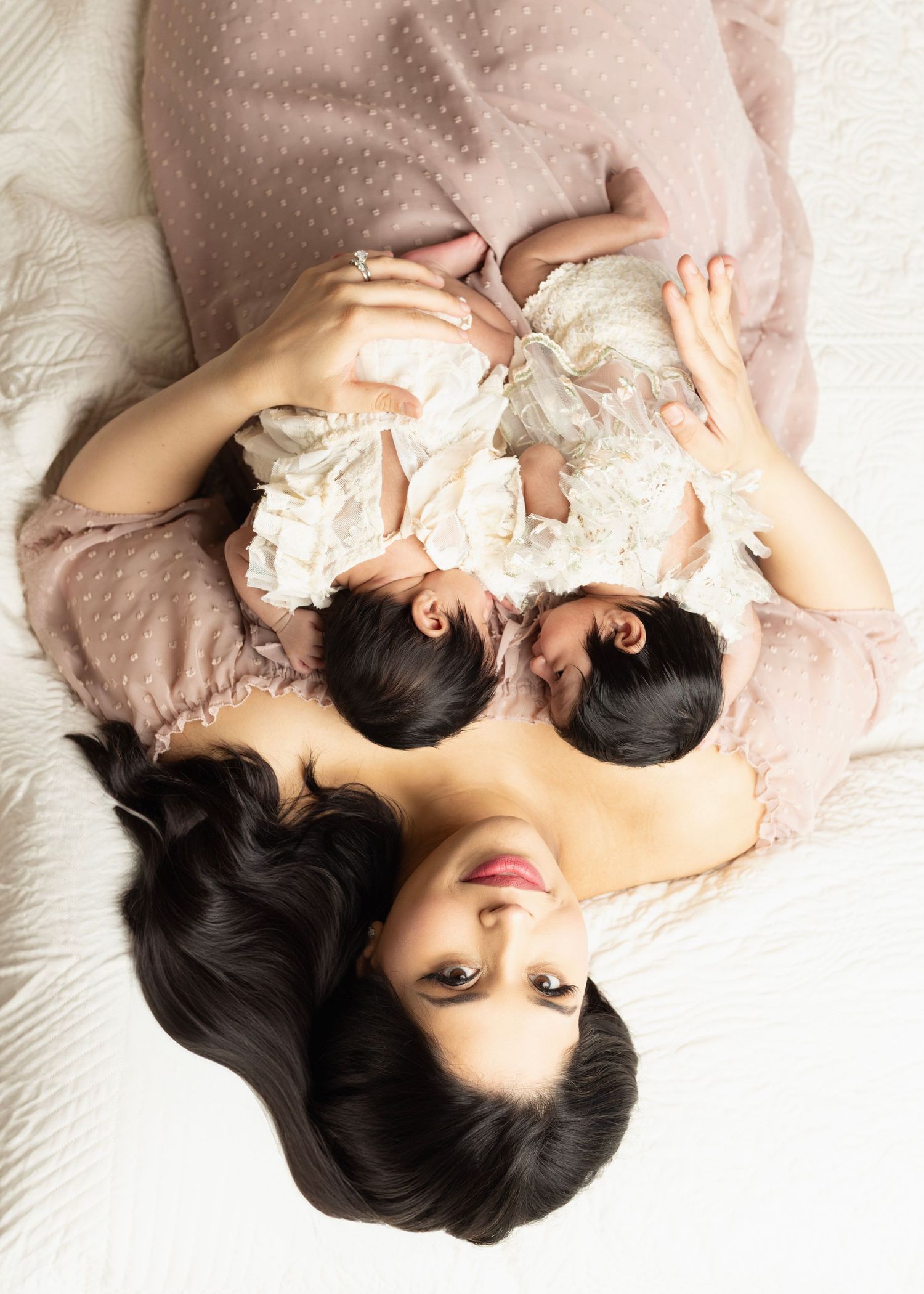 mom lays in bed with twin newborn girls resting on her chest sleeping peacefully. Mom looks deeply into the camera.