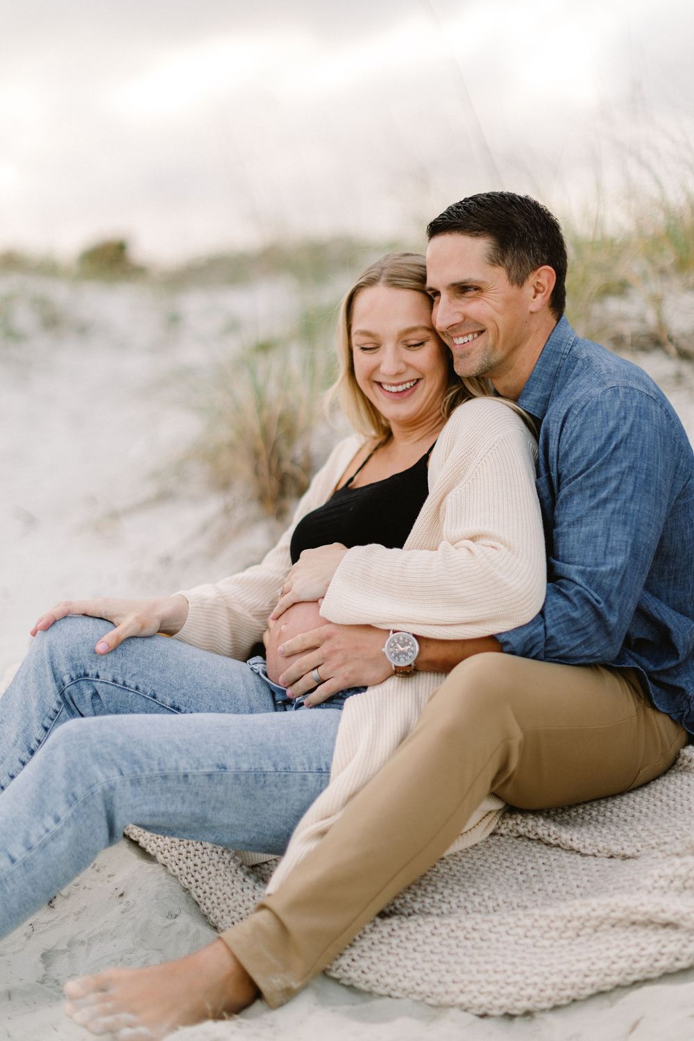 Relaxed maternity photo of couple sitting on blanket at beach.