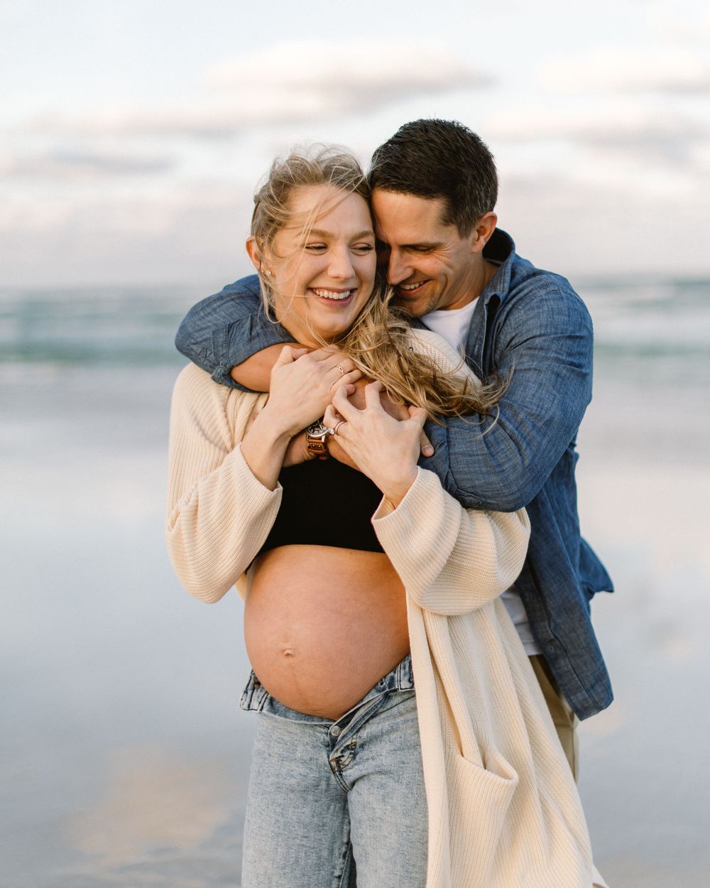 An expectant couple shares a joyful moment together on a misty beach.