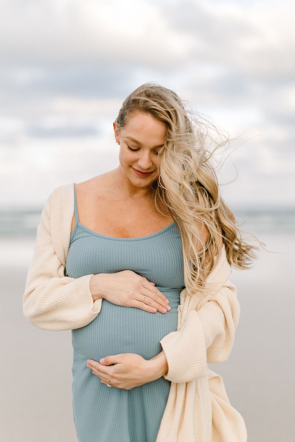 An expectant mother in a blue dress caresses her belly on a cloudy beach.