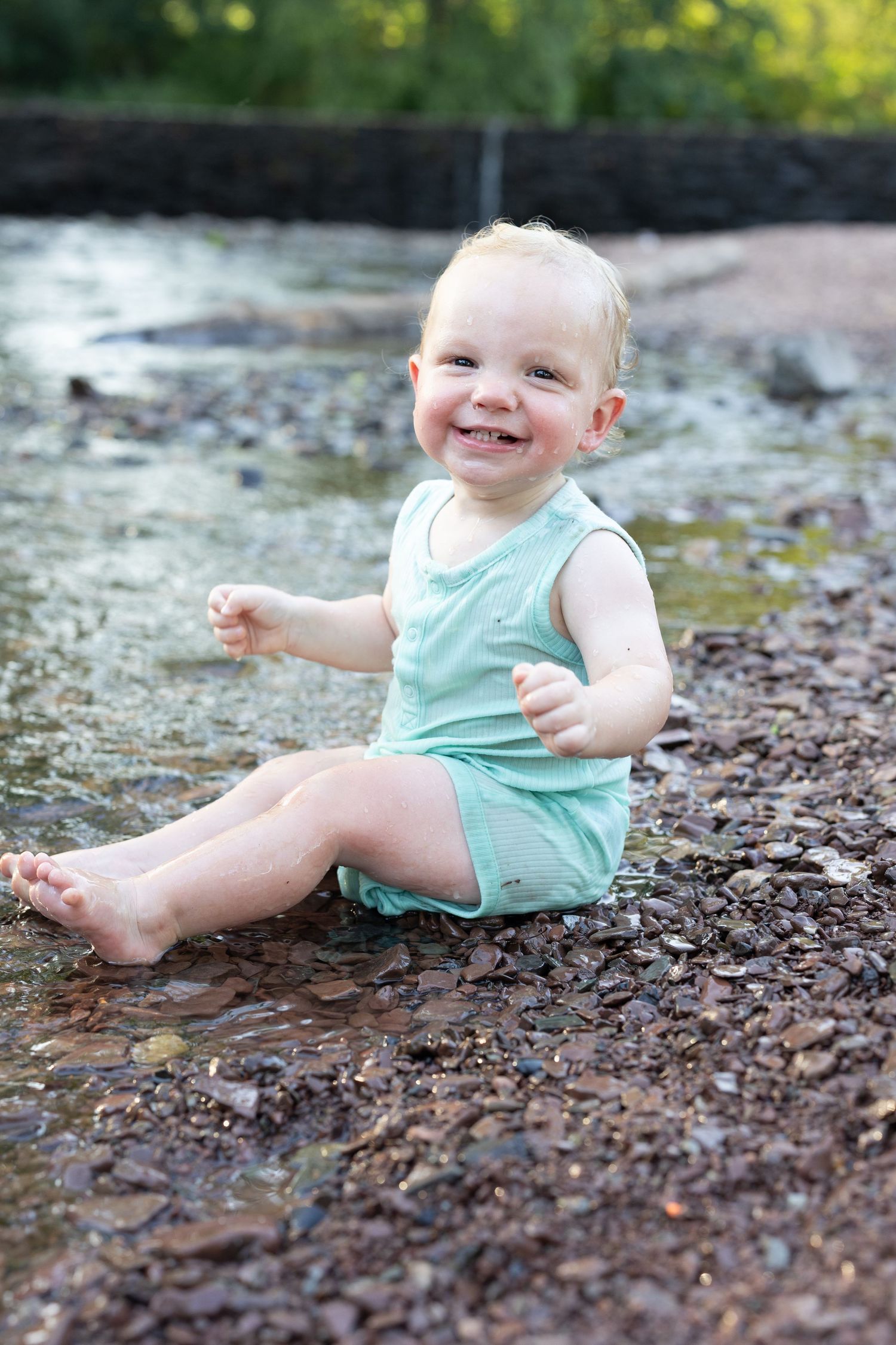 A happy baby in a mint green romper sits and plays on a rocky creek bed near flowing water.