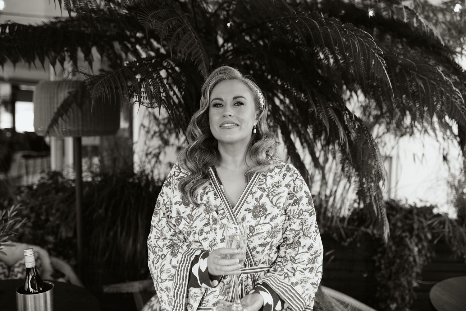 A vintage black and white photograph showing someone wearing a floral patterned robe while seated beneath tropical palm fronds.