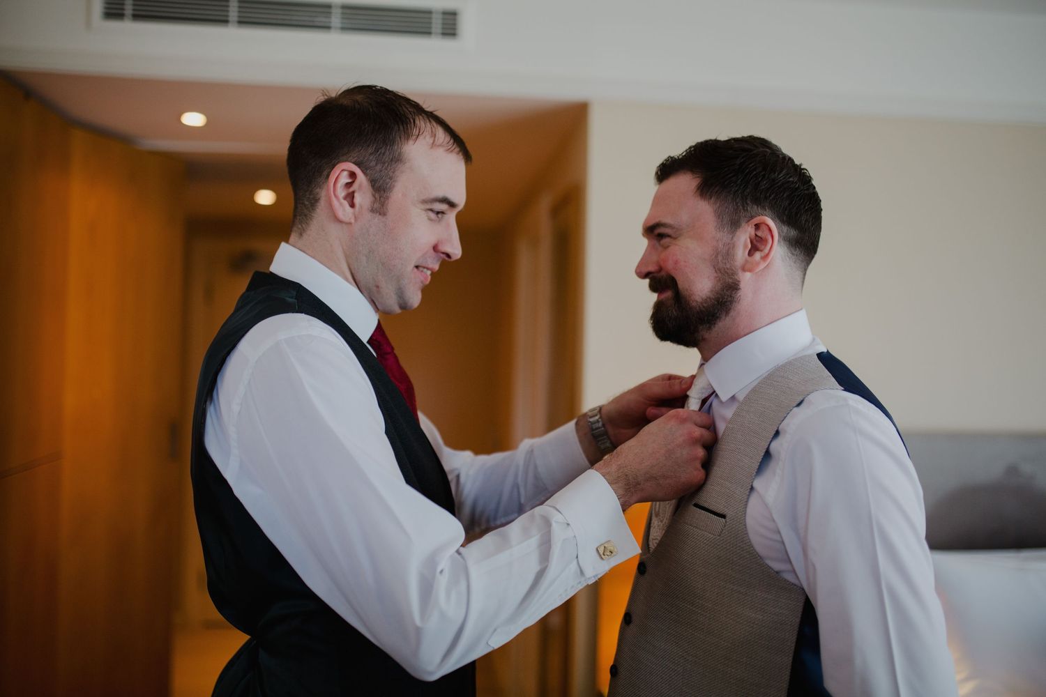 Two men in white dress shirts help each other with formal attire preparations during a wedding day.