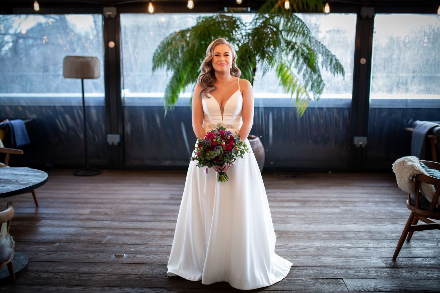 An elegant bride in a flowing white gown holds a vibrant bouquet while standing in a stylish indoor venue with palm decor.