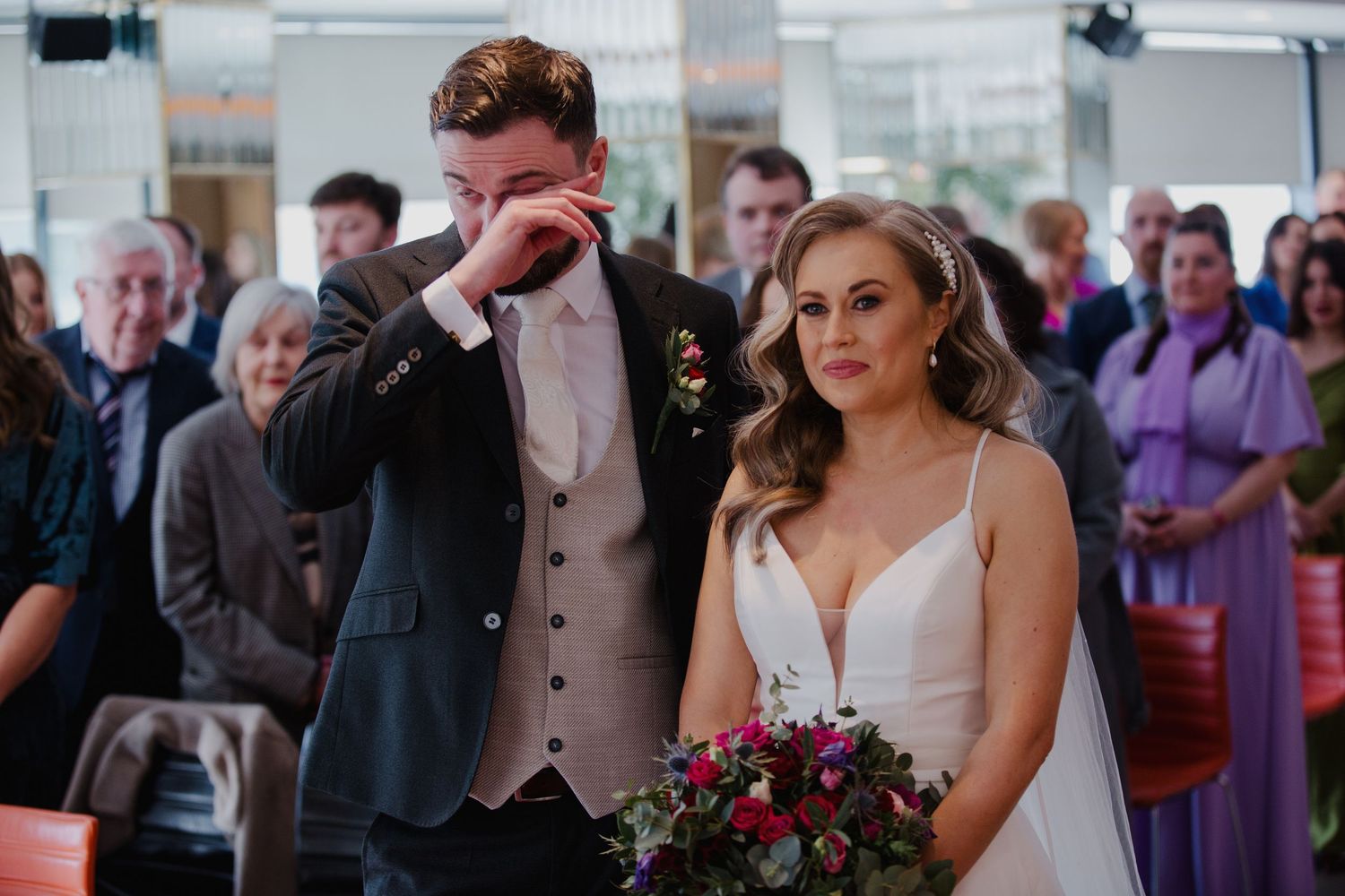 A couple shares an emotional moment during their wedding ceremony with guests watching and a red bouquet in hand.