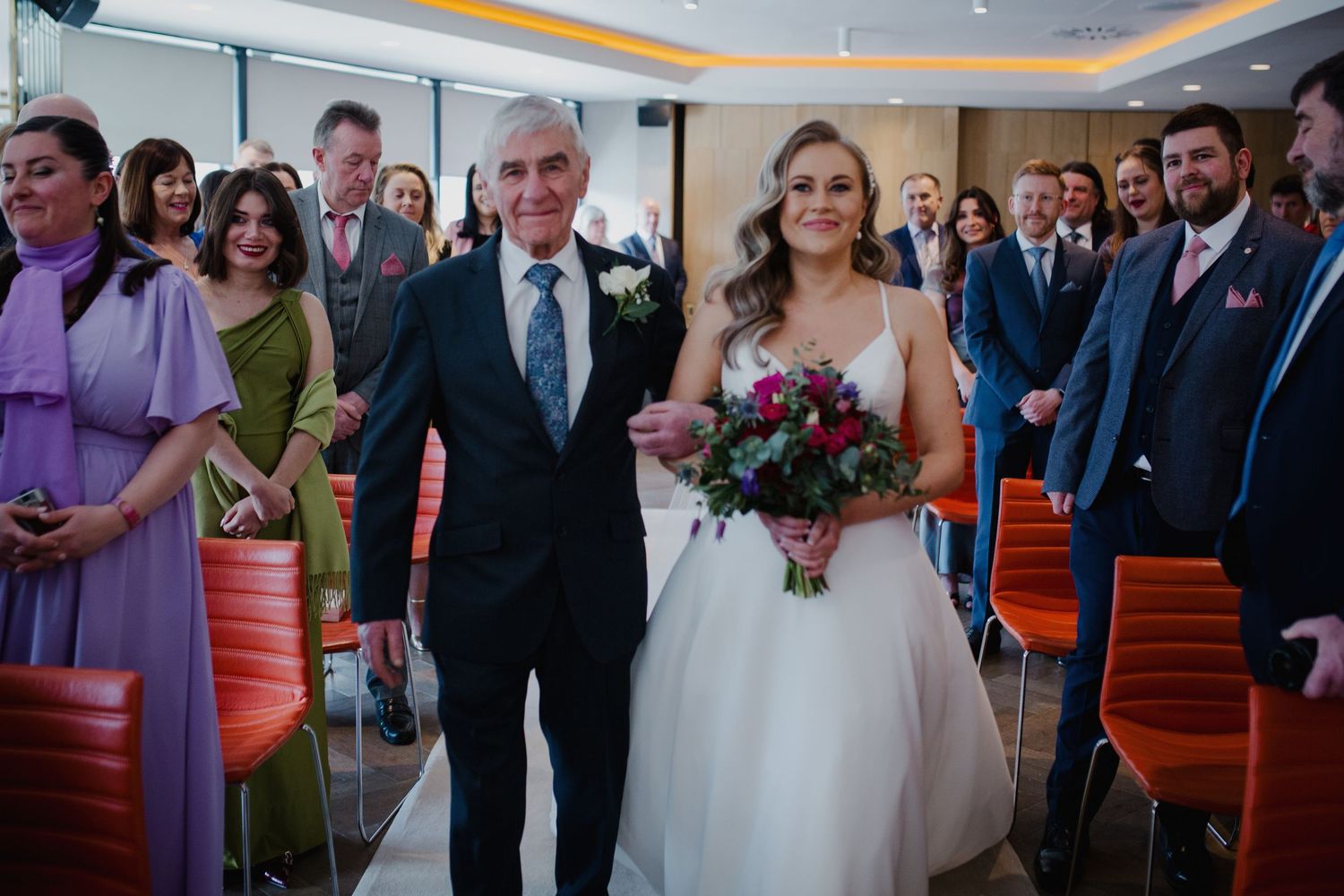 Wedding ceremony scene showing a bride in a white gown and bouquet walking down the aisle in an indoor venue.