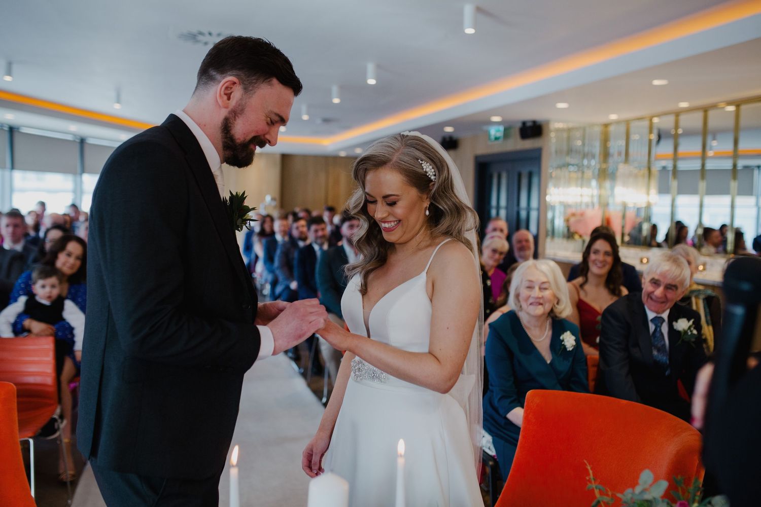A joyful moment during a wedding ceremony as a couple exchanges rings while guests look on smiling in the background.