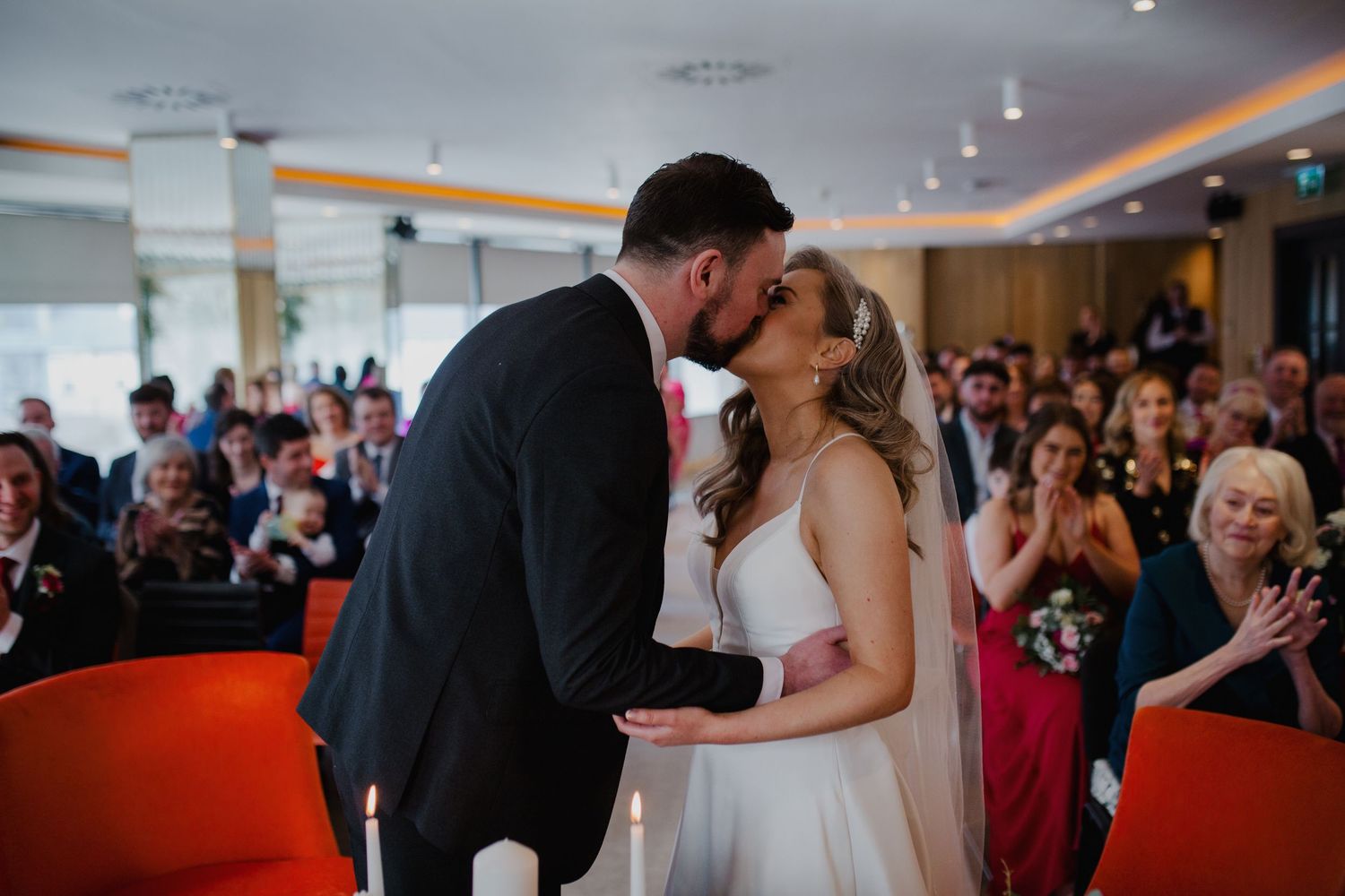 A couple shares their first kiss as newlyweds while wedding guests applaud in a modern venue with red decor.