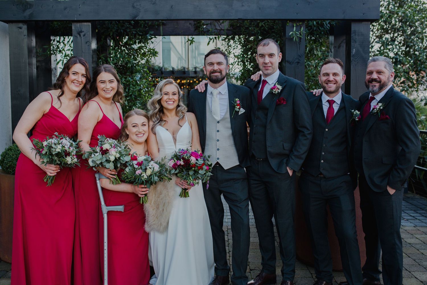 Wedding party poses outdoors wearing red bridesmaids dresses and black suits with red accents.