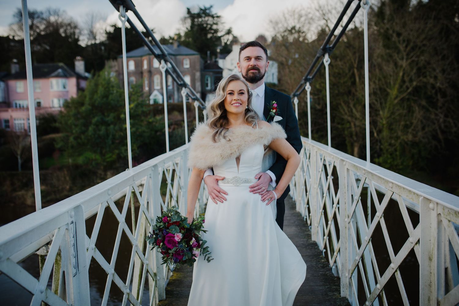 A couple poses on a white iron bridge with pink houses visible in the background during their wedding.