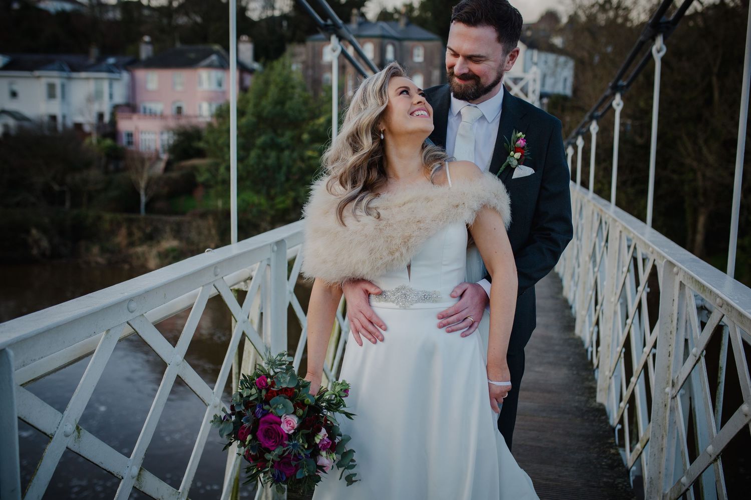 Newlywed couple share a romantic moment on white bridge at sunset holding vibrant purple floral bouquet.