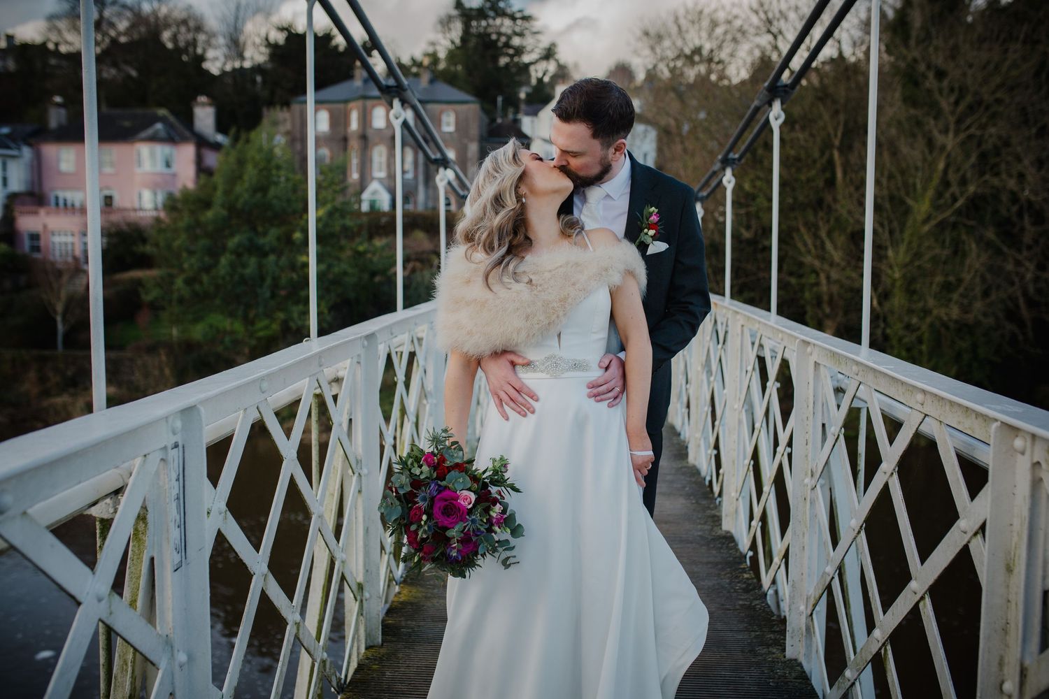 A romantic couple in wedding attire embrace on a white metal bridge while holding a vibrant purple bouquet.