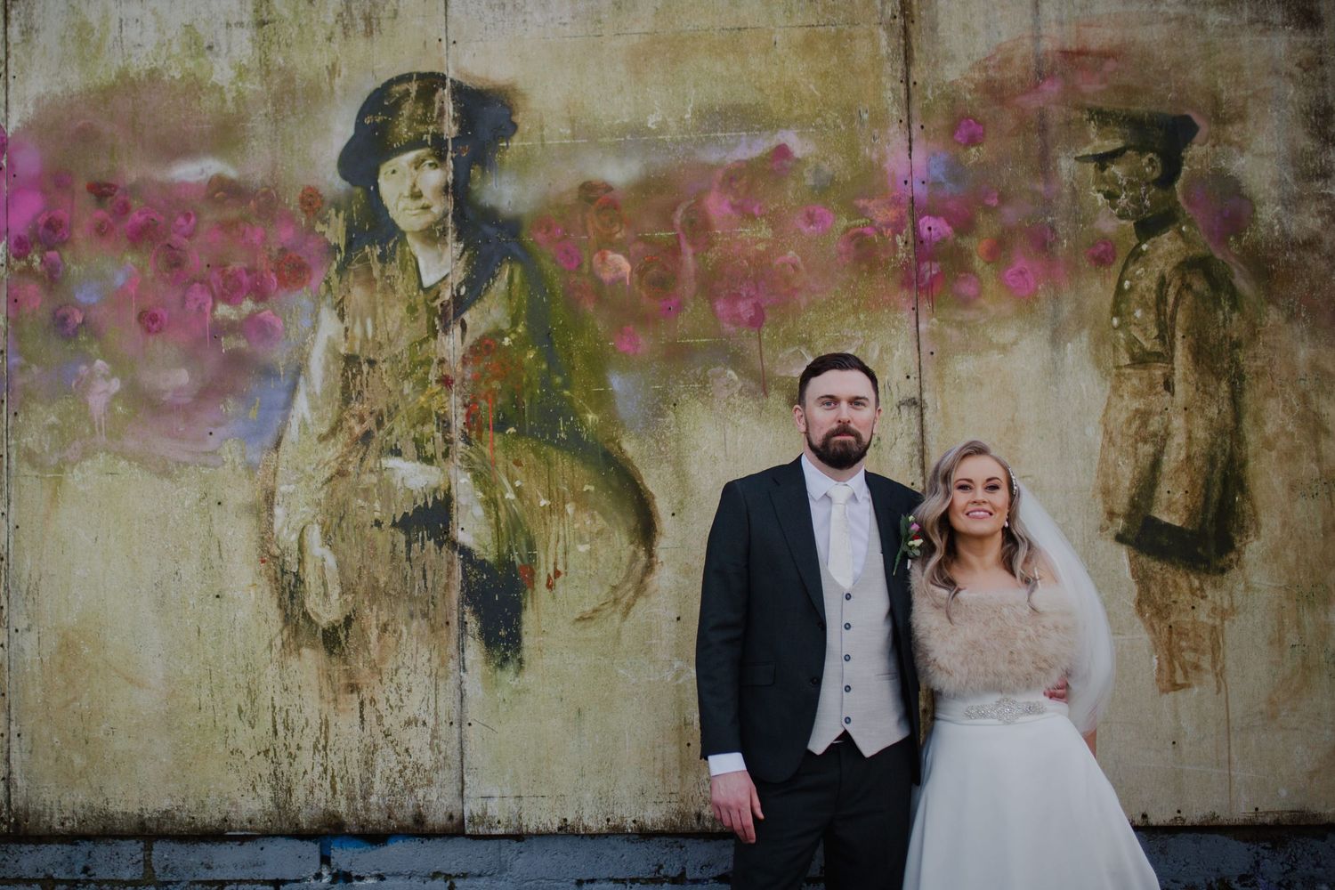 Wedding couple poses in front of distressed pink and beige textured wall creating artistic contrast.