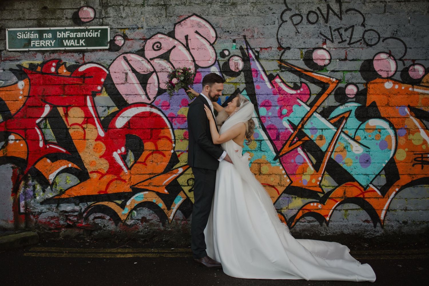 A couple in wedding attire embraces against a vibrant urban graffiti wall featuring colorful street art in shades of red and orange.