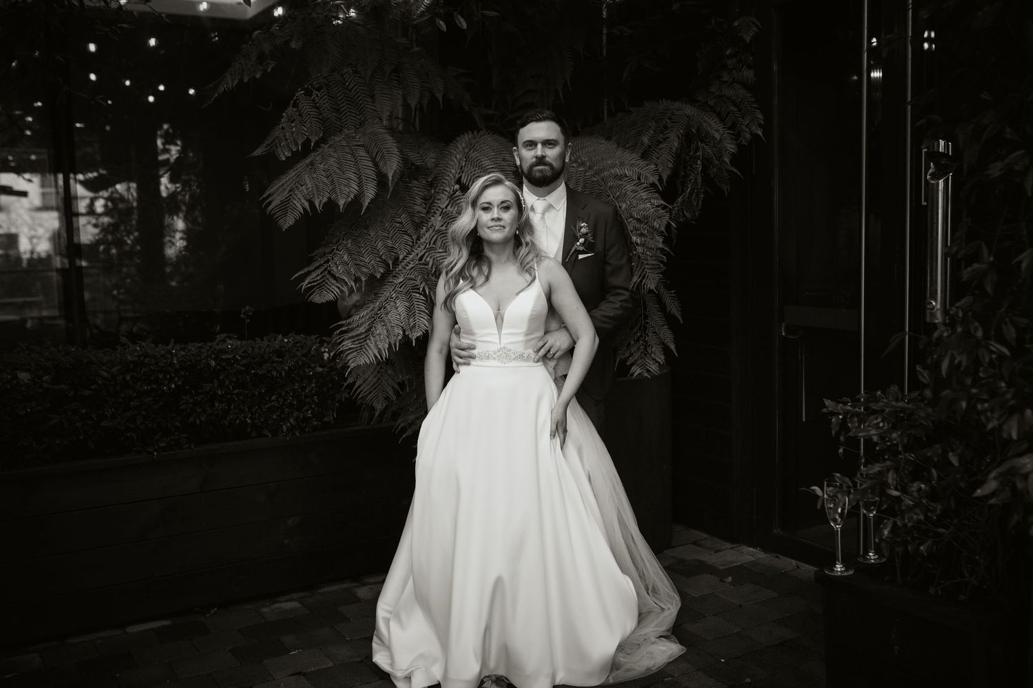 Black and white portrait of couple in formal wedding attire posing in front of tropical greenery.