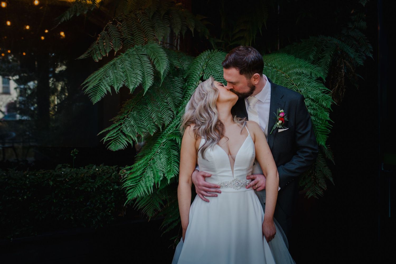 An intimate wedding portrait features a couple against dark green foliage in moody lighting.