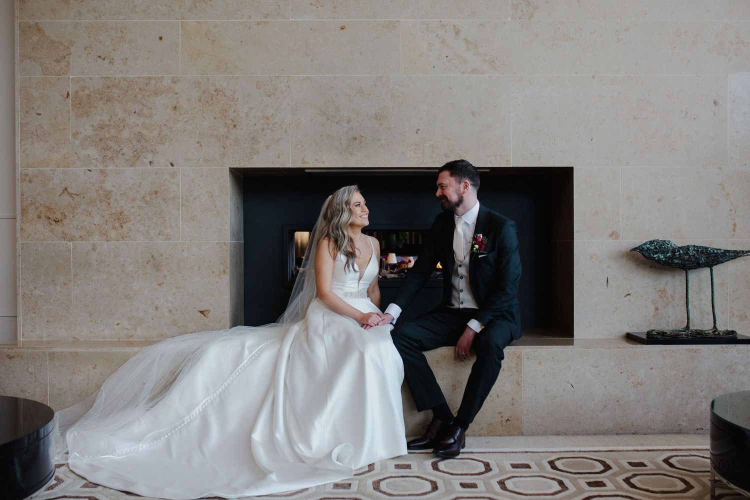 Couple in formal wedding attire share a private moment in front of a modern fireplace.