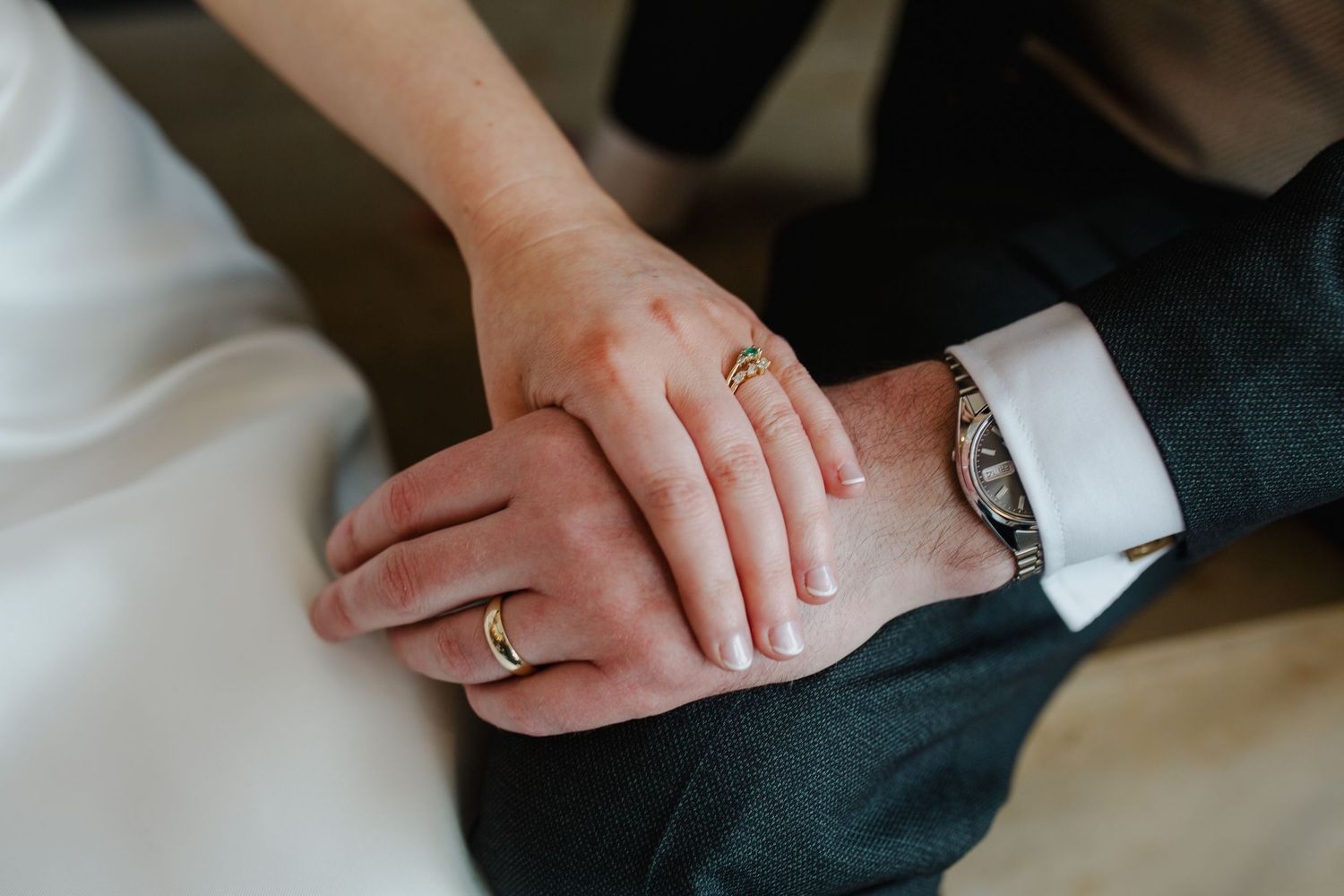 Close-up view of hands wearing wedding rings and a formal watch against dark and light clothing.