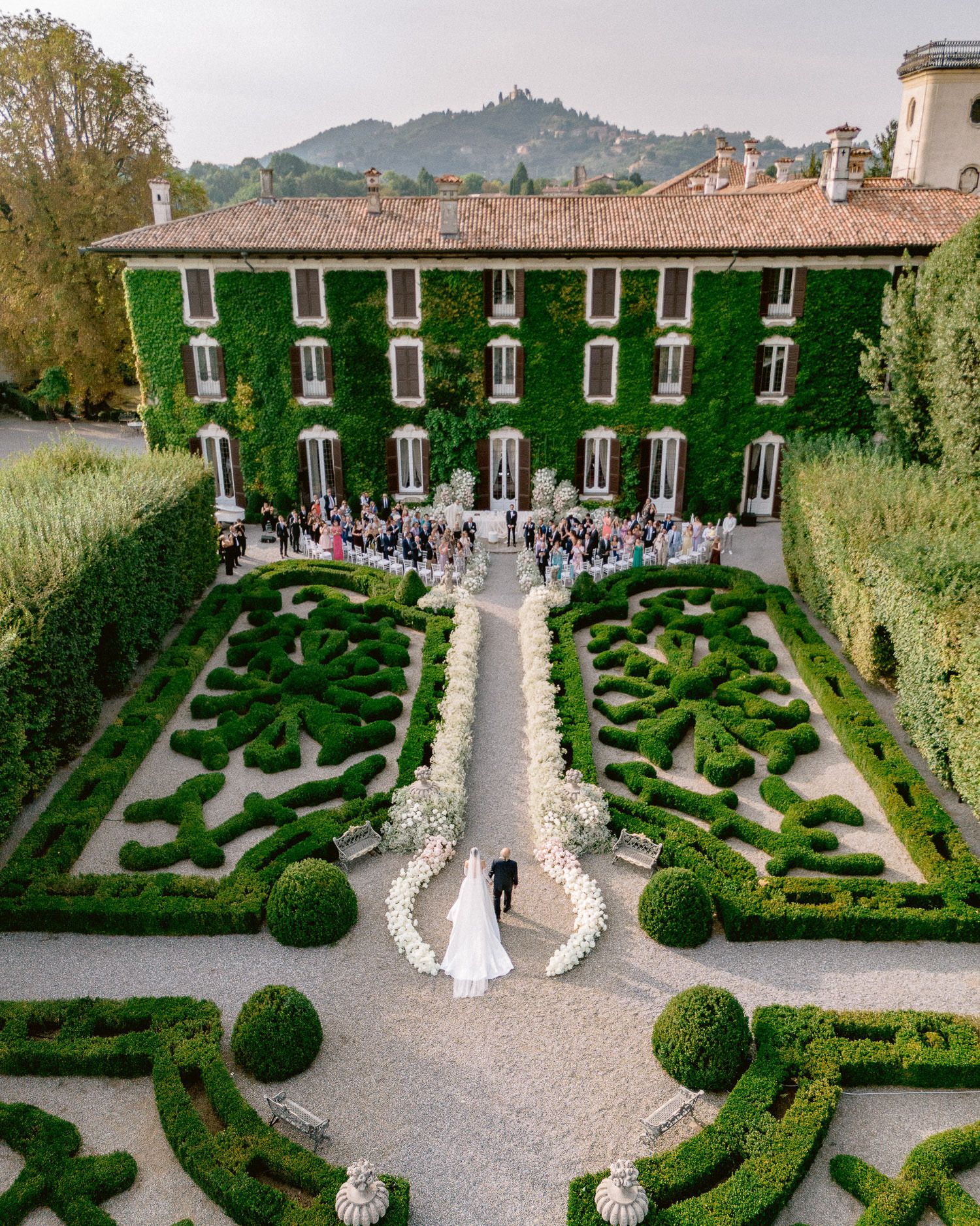 Aerial view of an Italian villa with elegant formal gardens featuring symmetrical hedges and a grand gravel pathway leading to the entrance.