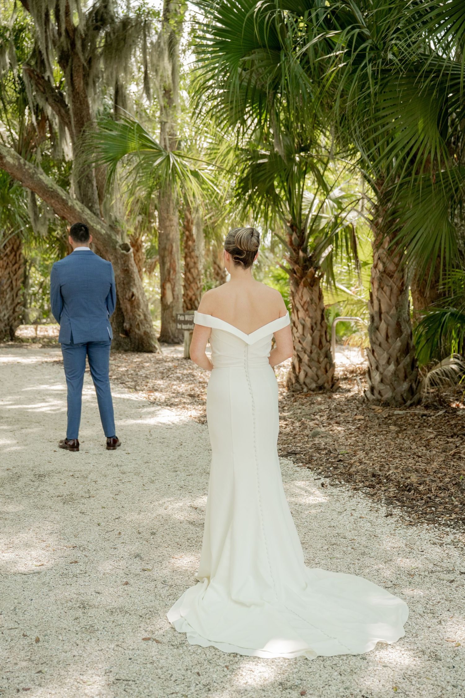 First look photos of couple in elegant wedding attire under palm trees and oak trees on a sandy path.