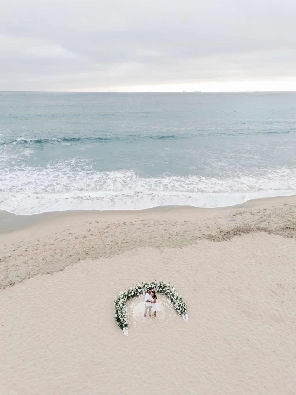 A romantic beach wedding ceremony setup with white floral archway and chairs facing the ocean on a misty day.