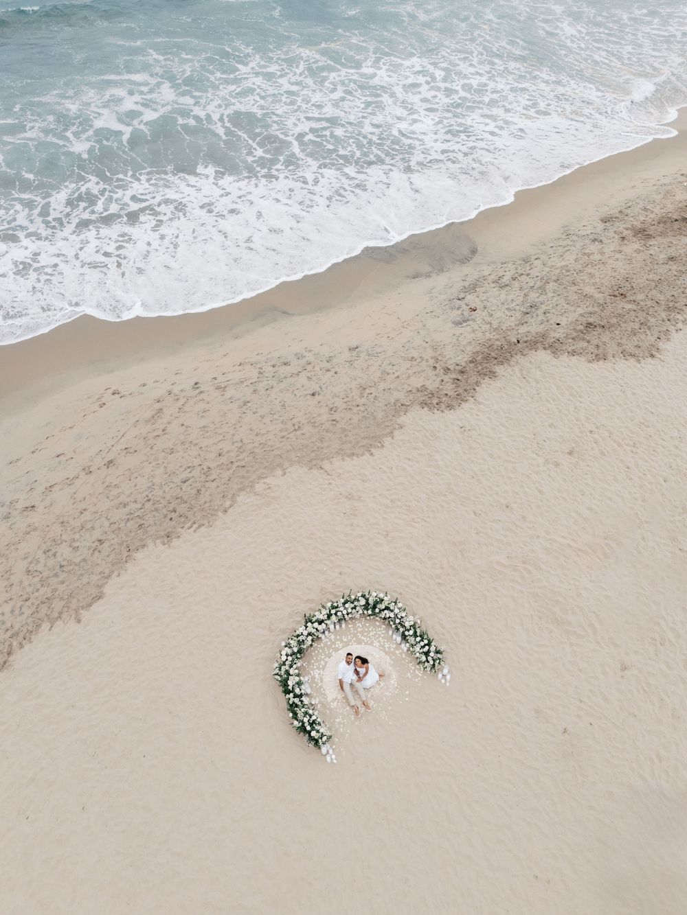 Aerial view of a couple lying in a crescent-shaped floral arrangement on a sandy beach with turquoise waves.