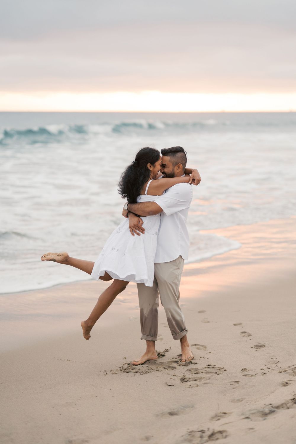 Two people in white outfits dance together romantically at sunset on a sandy beach with ocean waves in the background.