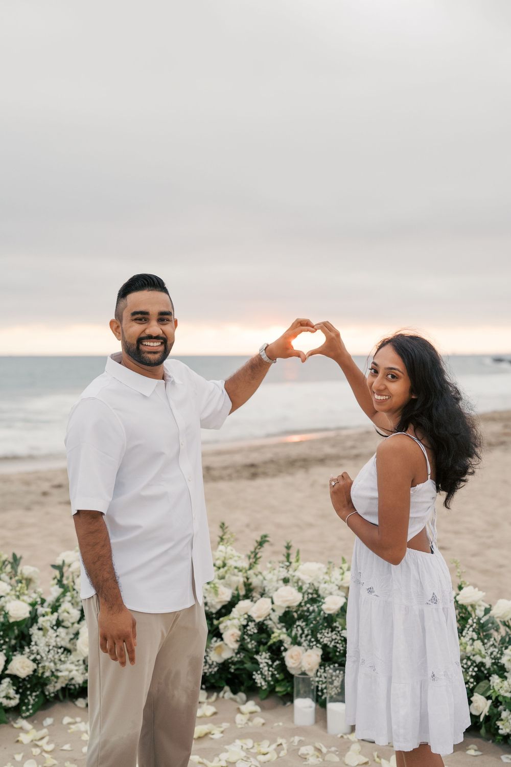 A couple shares romantic moments on the beach at sunset with white floral decorations arranged in the foreground.