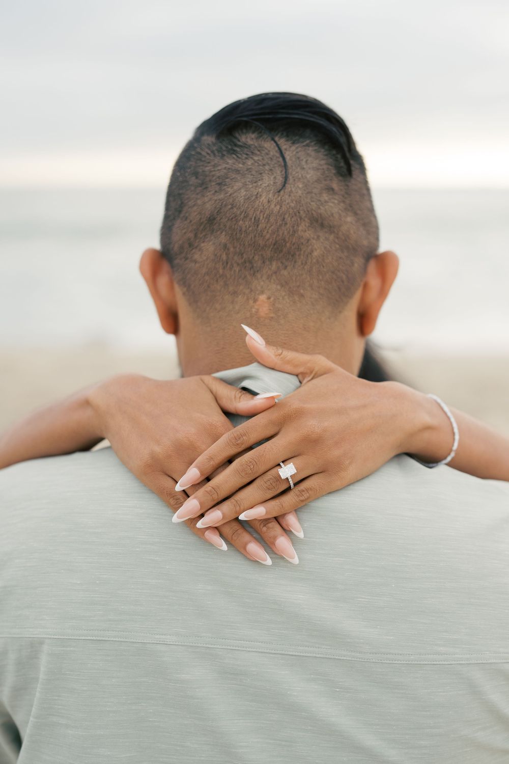 Arms wrapped around someone's back from behind, showing a wedding ring on their manicured hand against a light gray shirt.