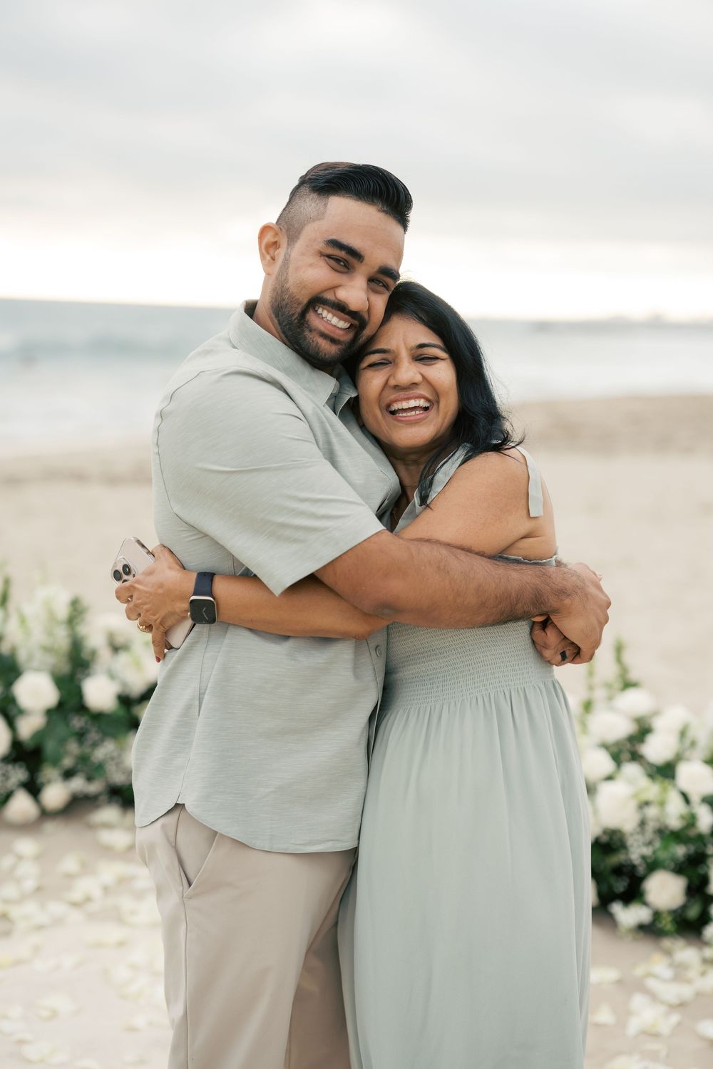 Happy couple embraces and laughs together on a beach with white flowers and ocean in background.
