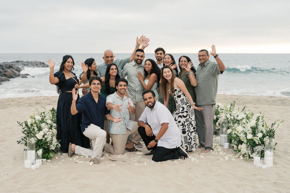 A happy group poses together on a beach at sunset with white floral arrangements decorating the sand.