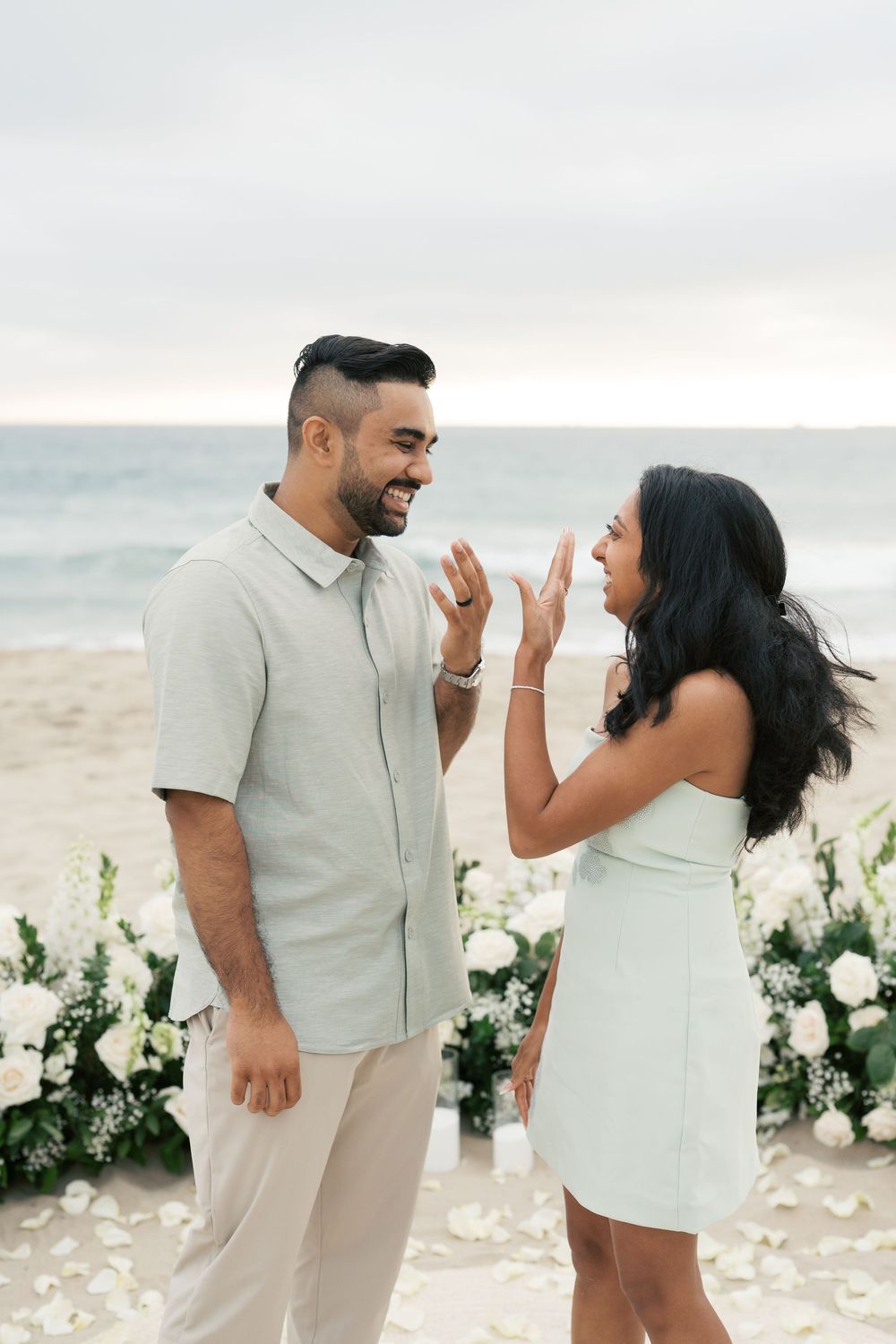 A joyful couple shares a candid moment together on a beach decorated with white flowers at sunset.