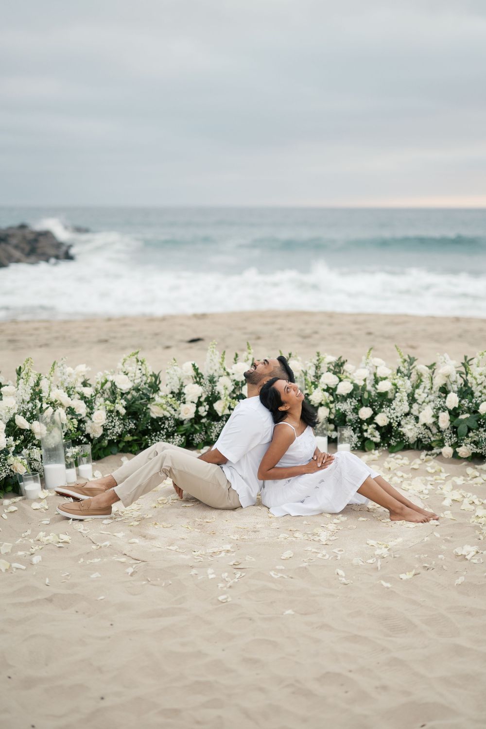A romantic beach photo series with a floral archway and couple laying in the sand by the ocean waves at sunset.