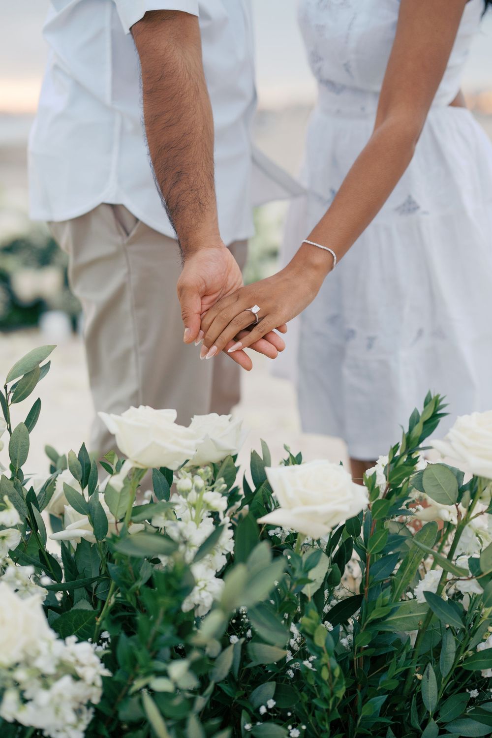A couple holds hands above an arrangement of white flowers and foliage at a wedding ceremony.