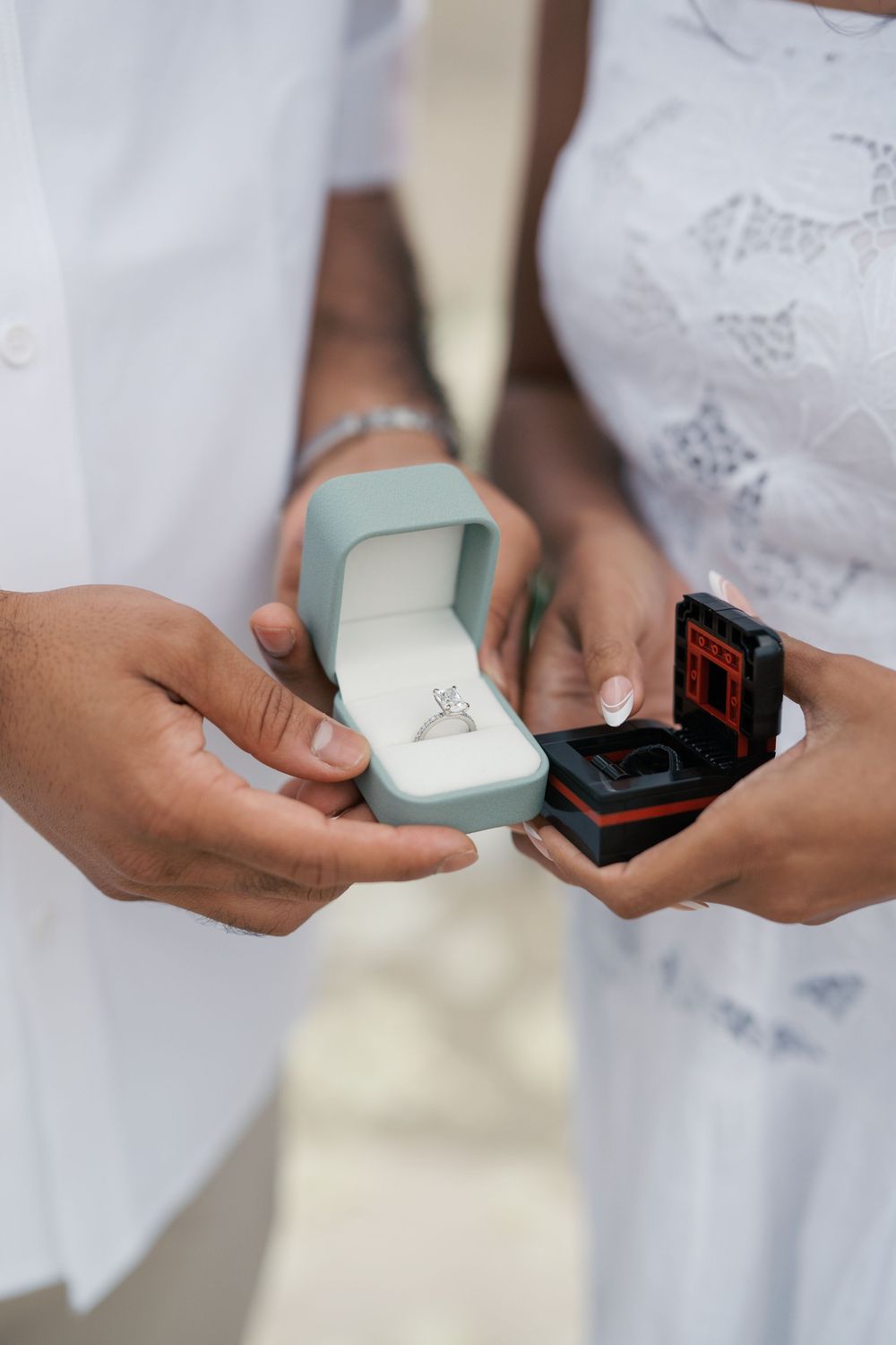 Two hands exchange small ring boxes in white formal attire during a special ceremony moment.