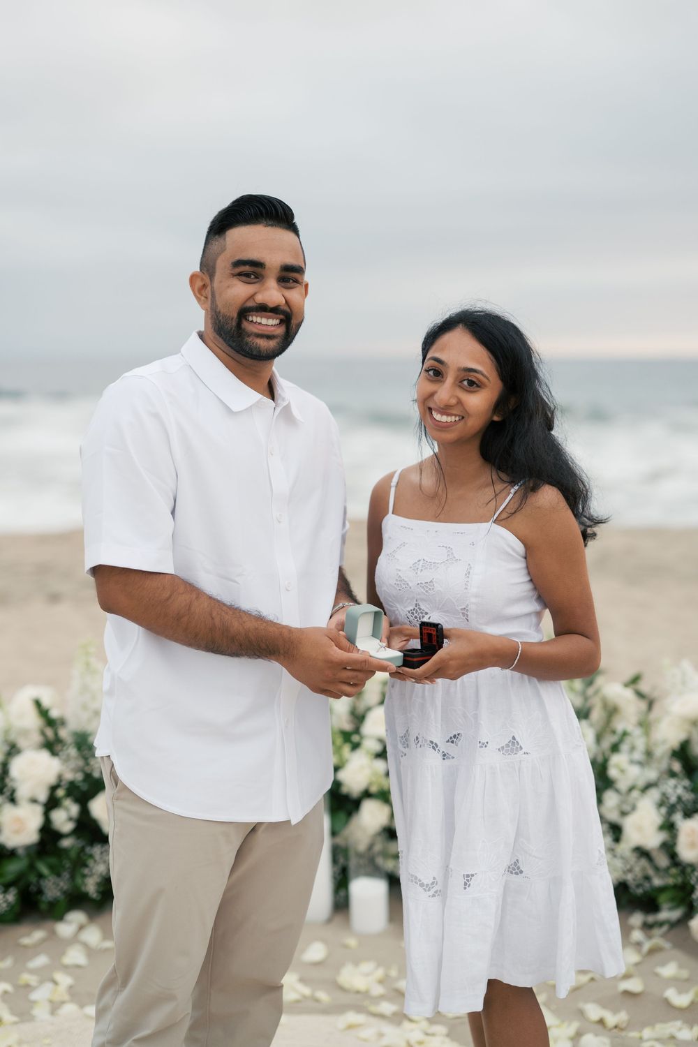 A couple in white outfits share a romantic moment during a beachside proposal with white floral decorations.