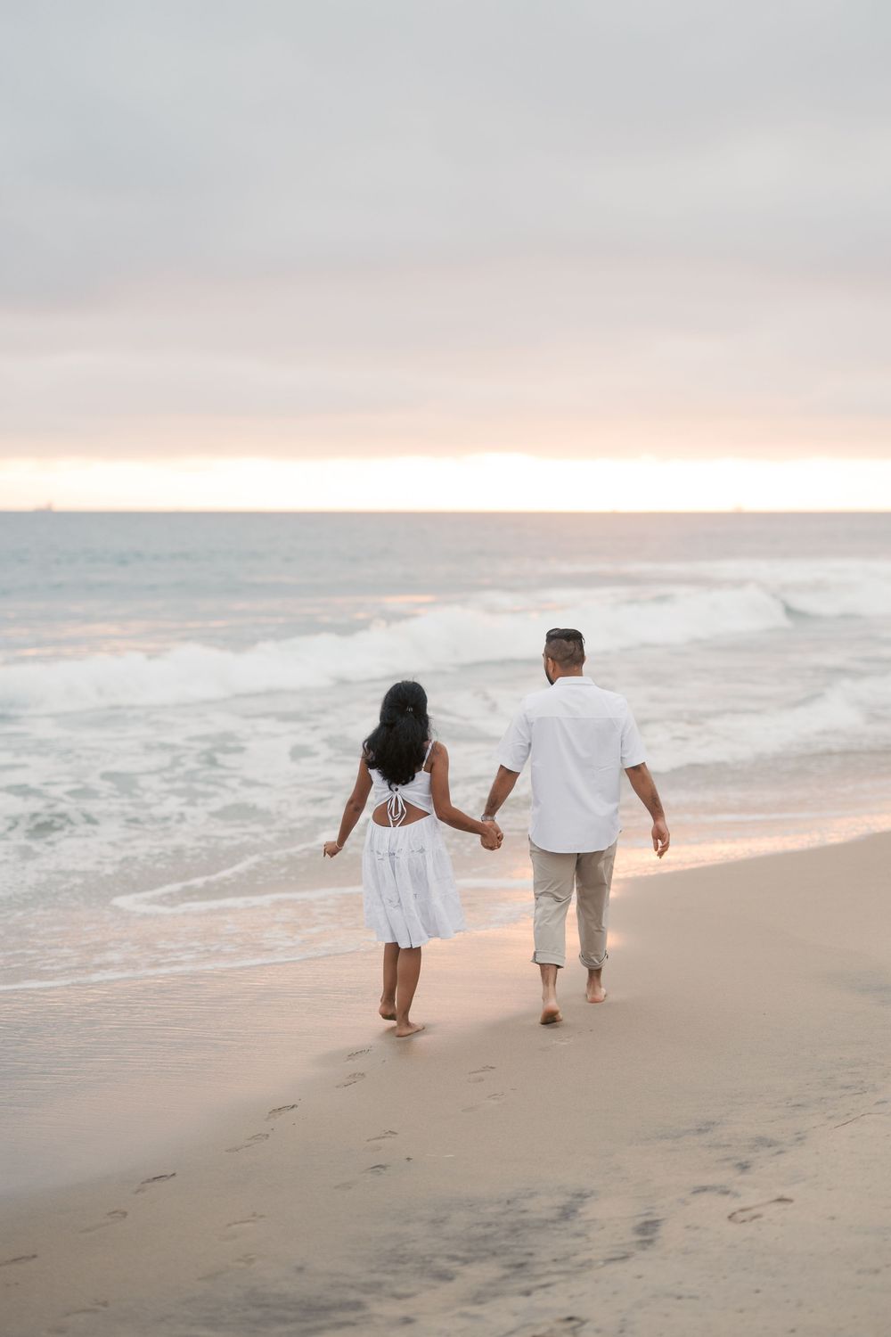 Couple walking hand in hand along a serene beach at sunset with waves crashing on the shore.