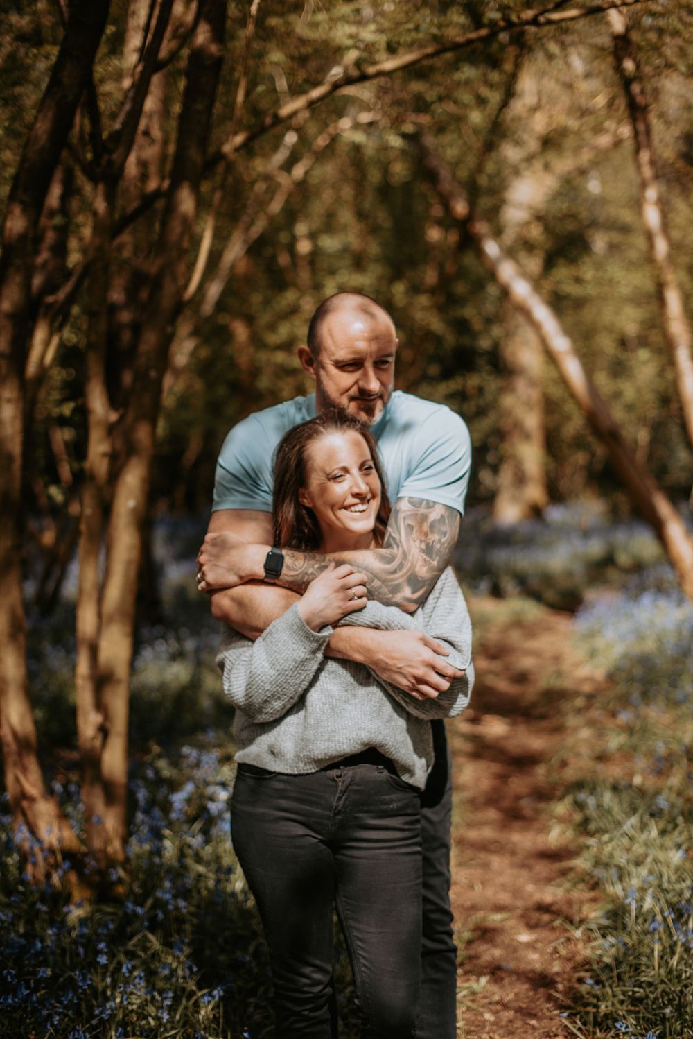 A couple embraces on a forest path surrounded by blooming bluebells and dappled sunlight filtering through trees.