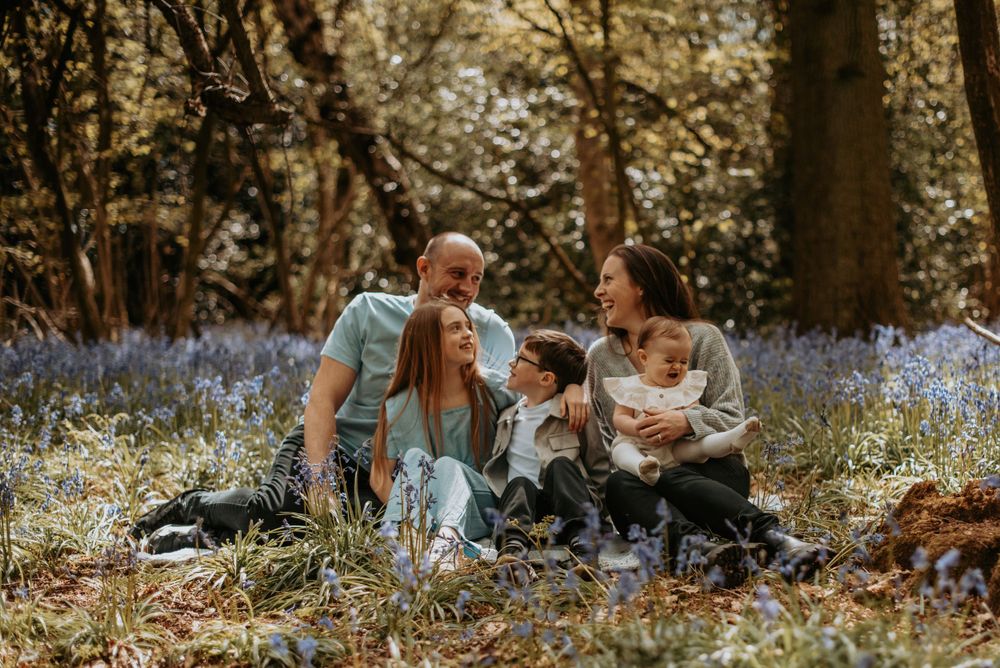 A family sits together on a blanket in a woodland filled with blooming bluebells on a sunny afternoon.