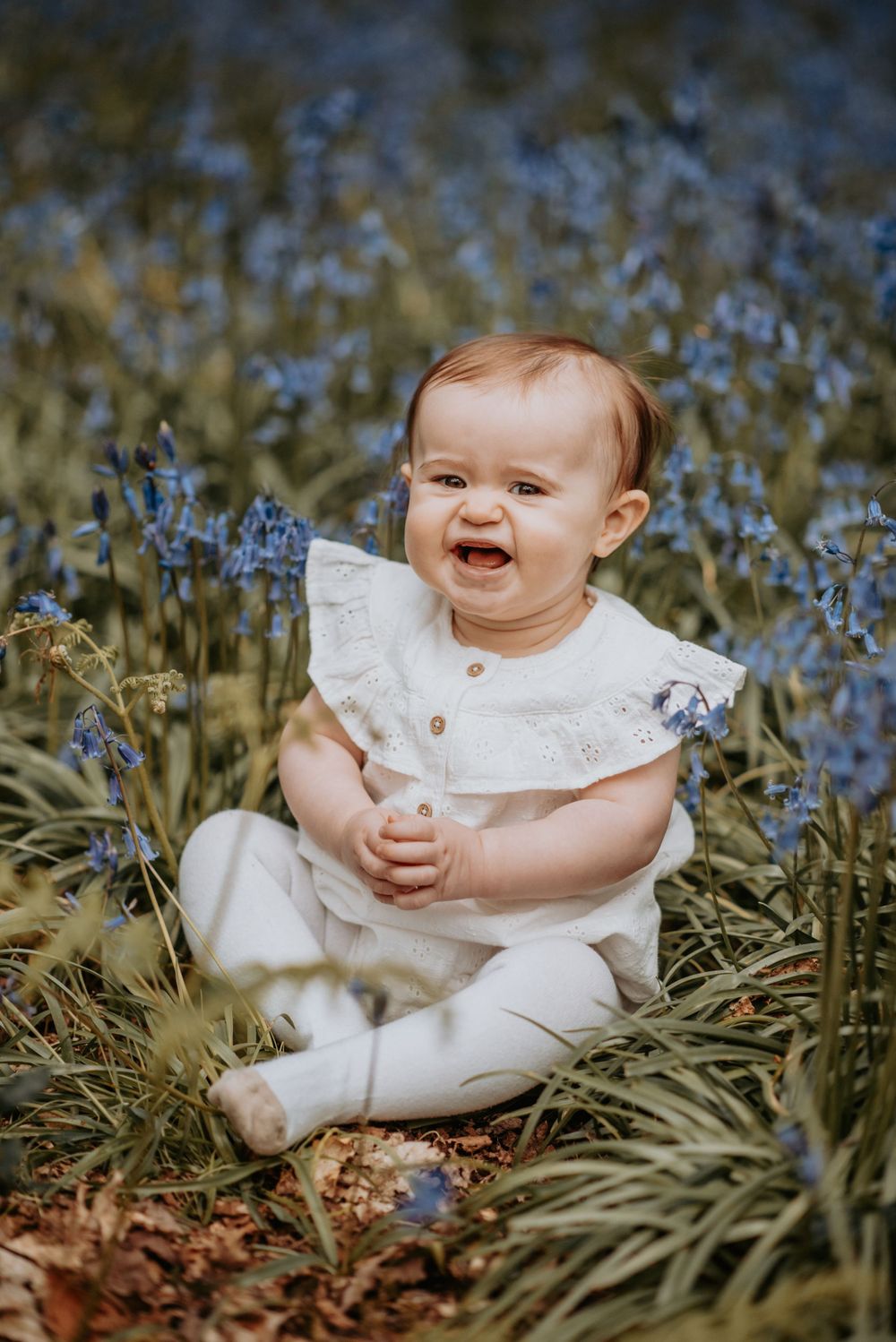 A smiling baby wearing white ruffle dress sits among blooming blue wildflowers in a sunny outdoor garden setting.