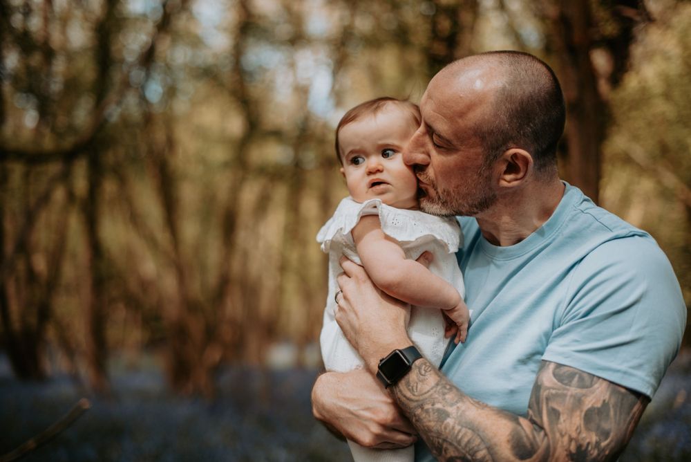 A tender moment shared between adult and baby in a light blue shirt against a blurred forest background.
