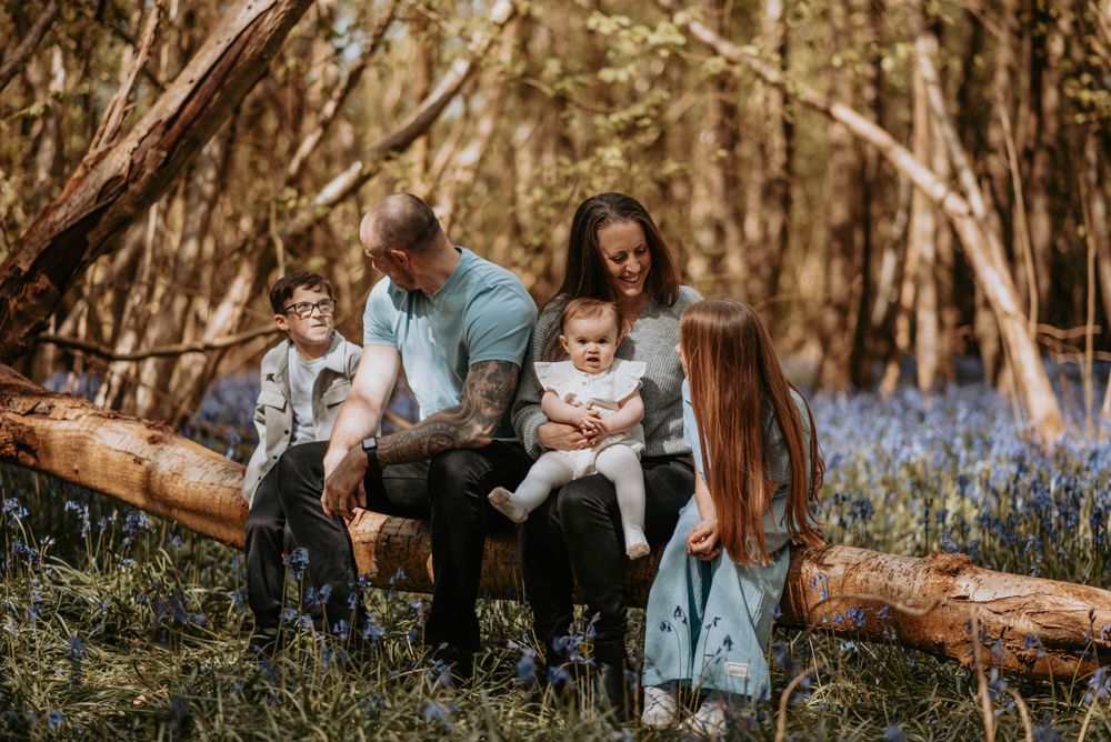 A family sits together on a fallen log in a forest filled with blooming bluebells during spring.