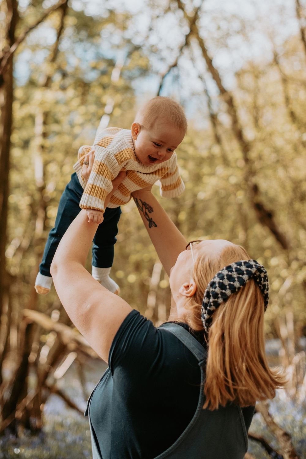A parent lifts a smiling baby wearing a striped sweater high into the air in an autumn forest setting.