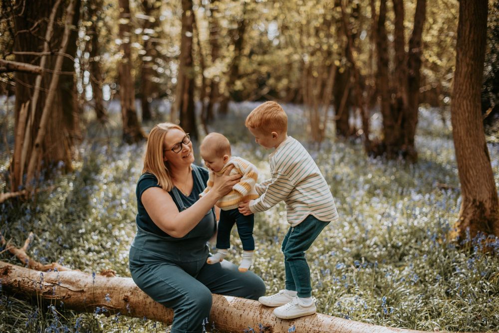 Family enjoying a tender moment together while sitting on a fallen log in a sunlit forest with bluebells blooming.