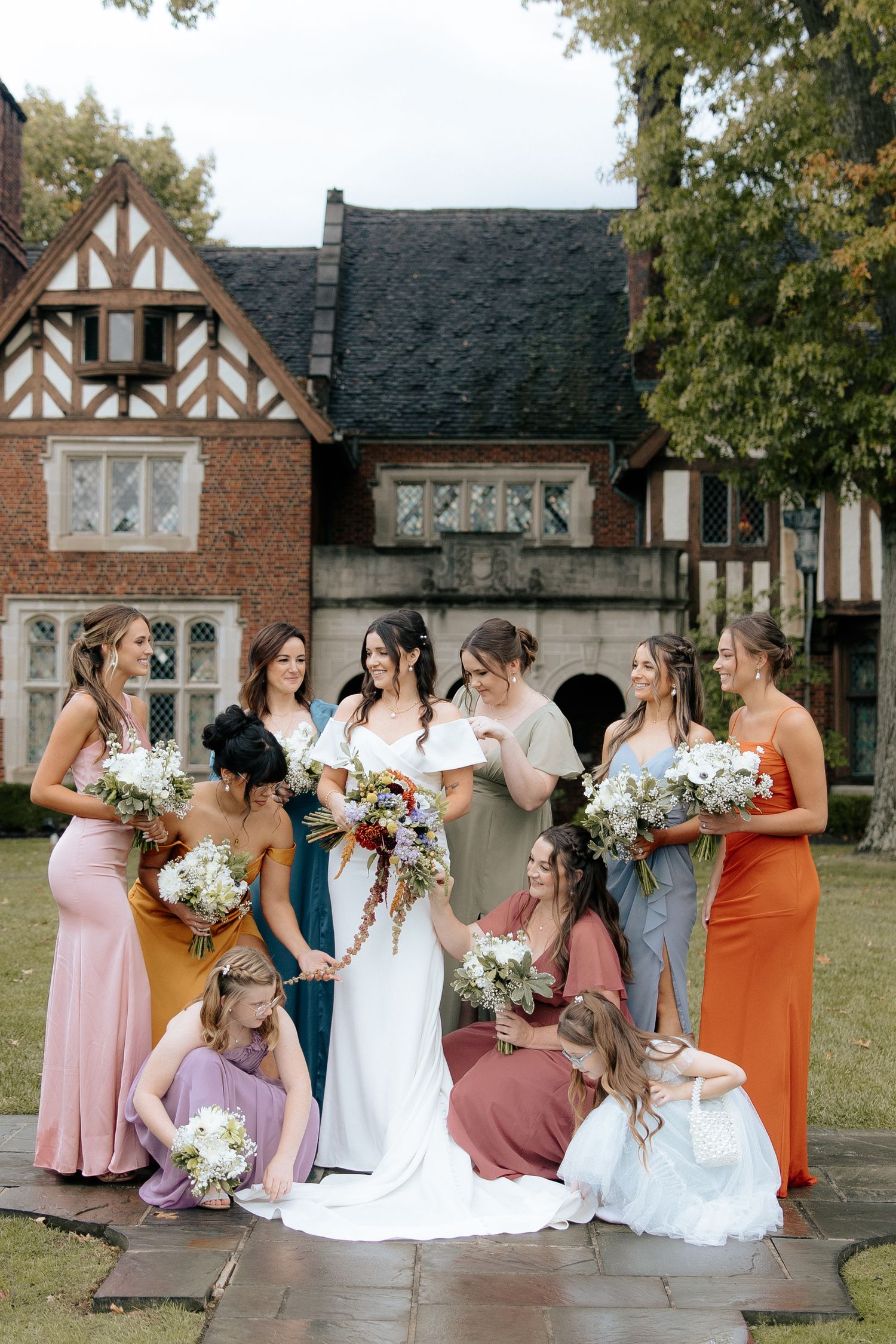 Bridesmaids in colorful dresses gather together with bouquets outside a Tudor-style brick mansion.