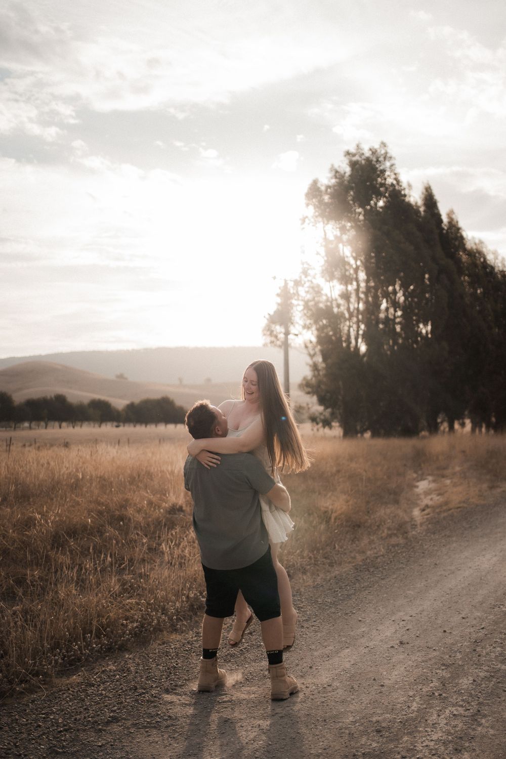 A couple embraces on a dirt road at sunset with golden fields and tall trees silhouetted against a dreamy sky.