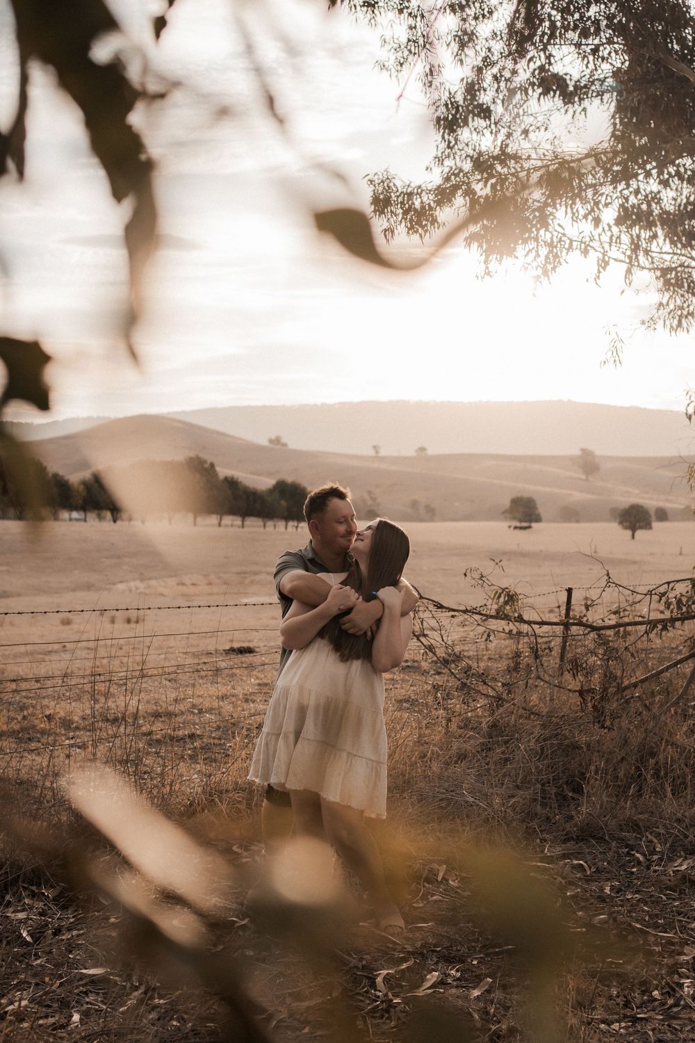 A romantic sepia-toned rural landscape scene with a couple embracing by a rustic fence at sunset.