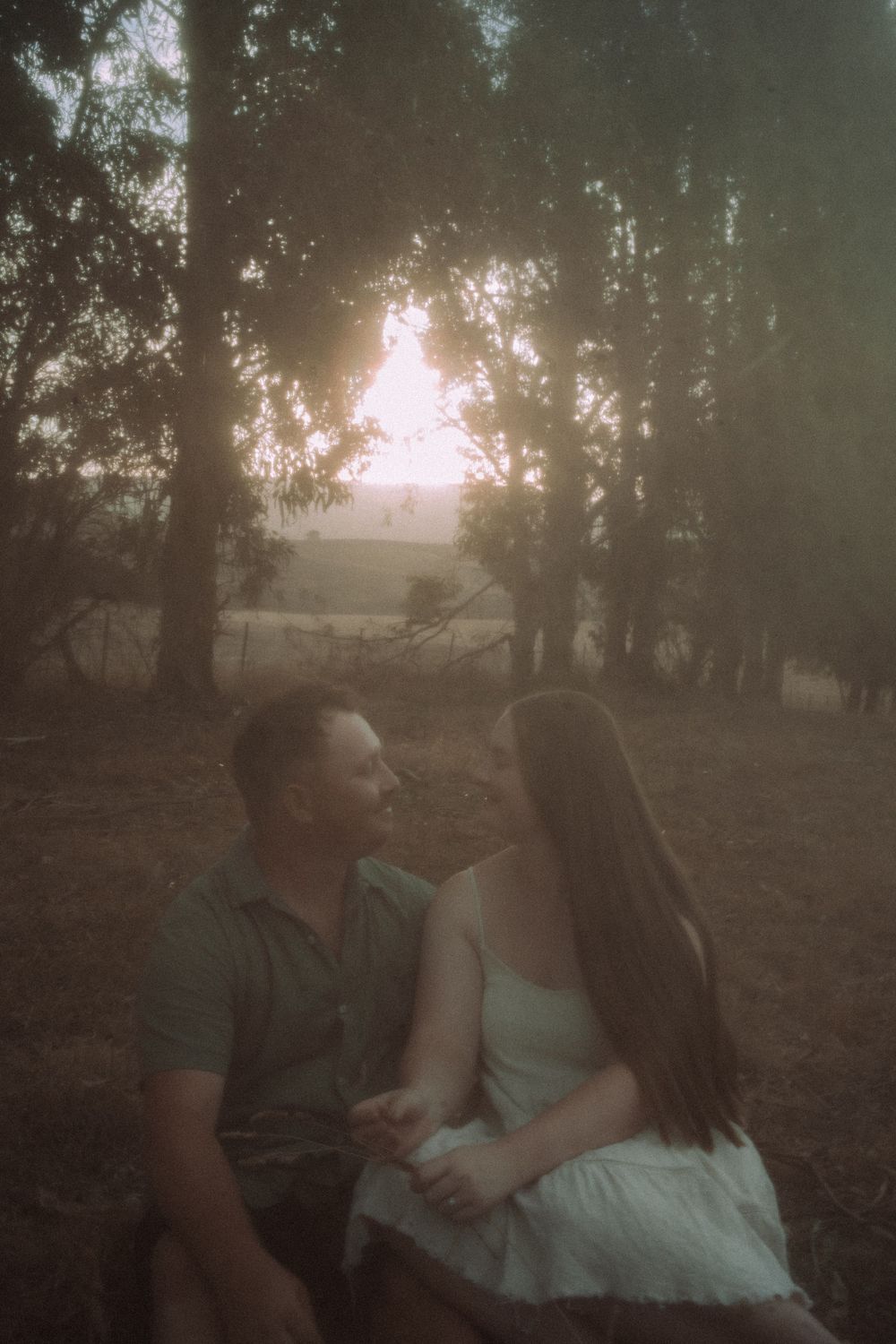 A romantic couple sits together in a dreamy forest setting at sunset with soft ethereal lighting and moody atmosphere.