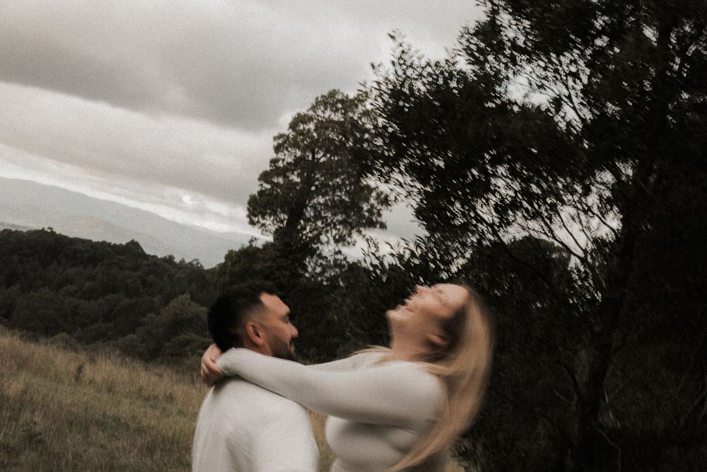Artistic black and white photo of a romantic couple embracing in a moody outdoor field at sunset.
